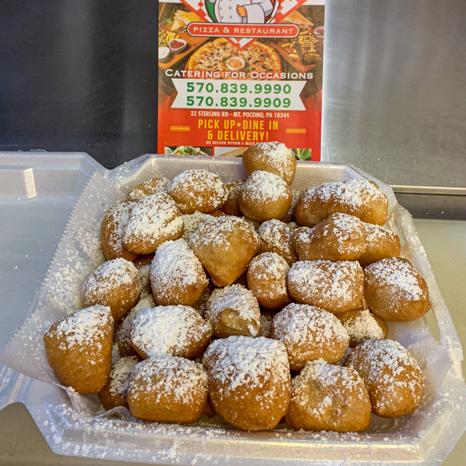 A white styrofoam container filled with golden-brown zeppole, generously dusted with powdered sugar.