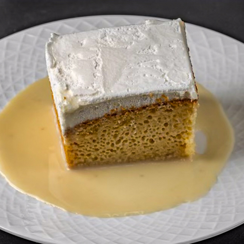 A slice of Tres Leches cake with white frosting sits in a pool of cream on a white plate.
