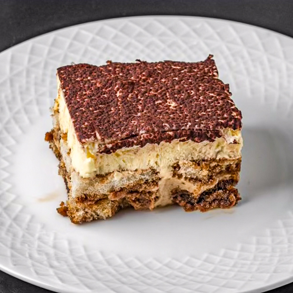 A delicious slice of tiramisu with cocoa powder on top, served on a white plate.