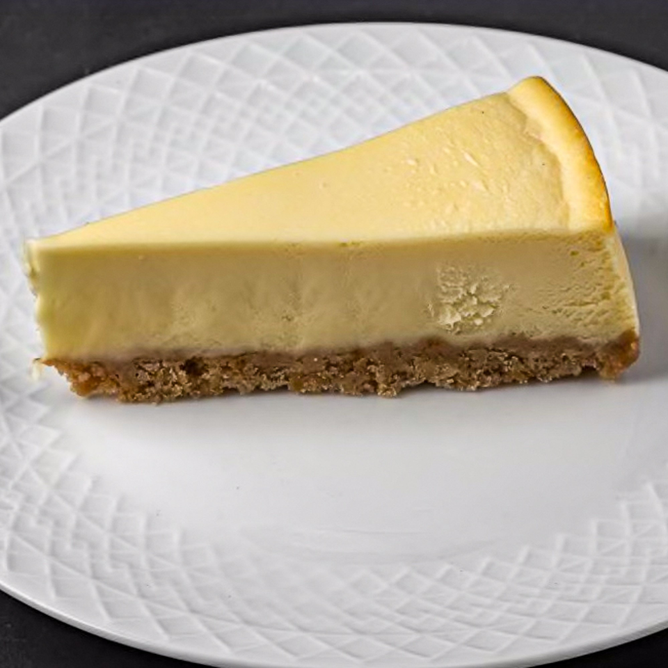 A slice of classic New York-style cheesecake with a graham cracker crust on a white plate.